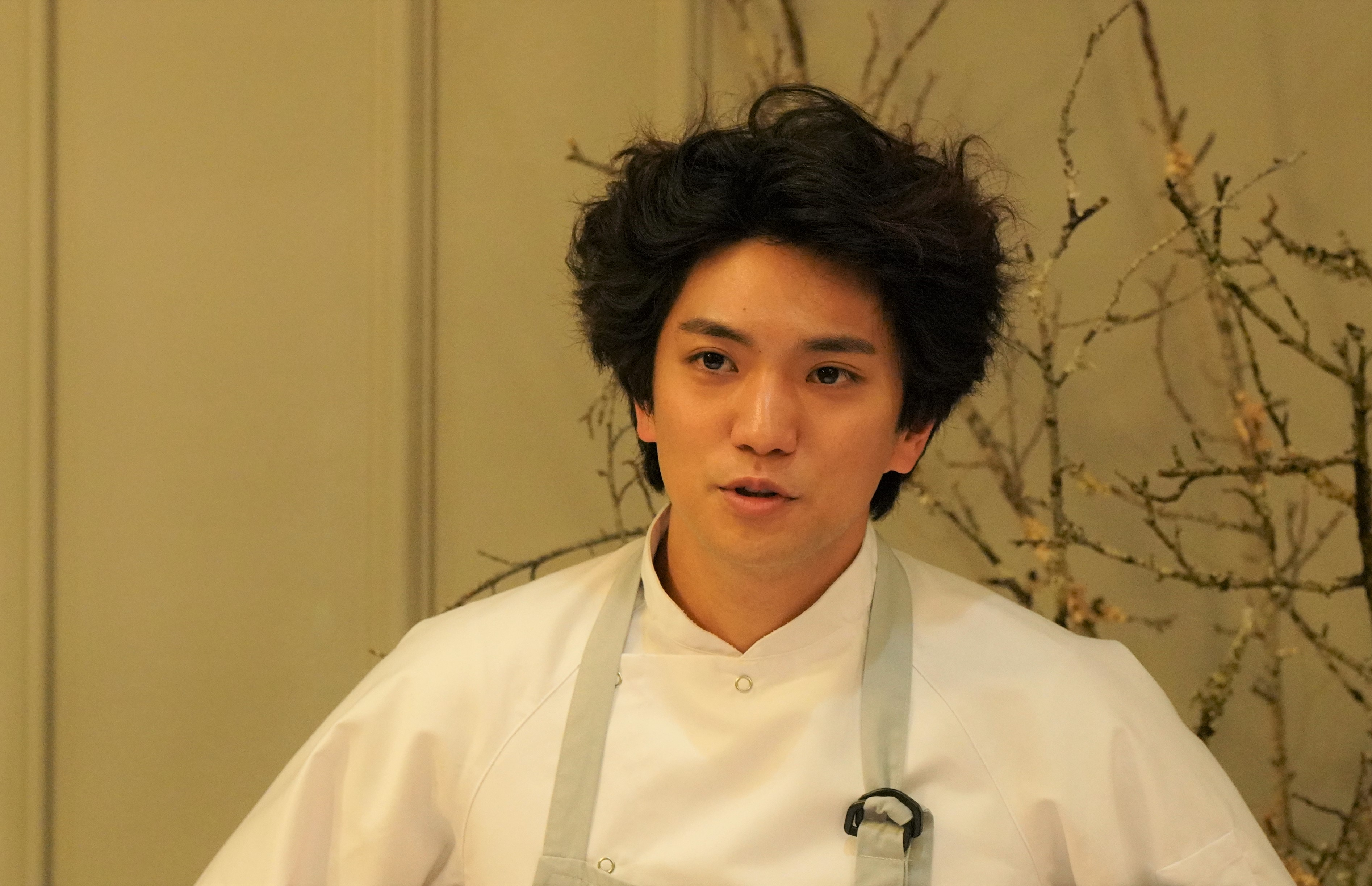 A young man in a chef's uniform speaks thoughtfully, framed against a warm, natural background.