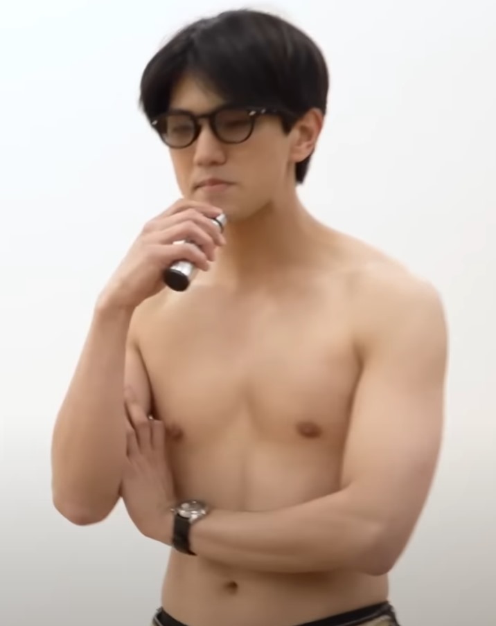 A shirtless young man holding a microphone, wearing glasses, and striking a thoughtful pose against a plain background.