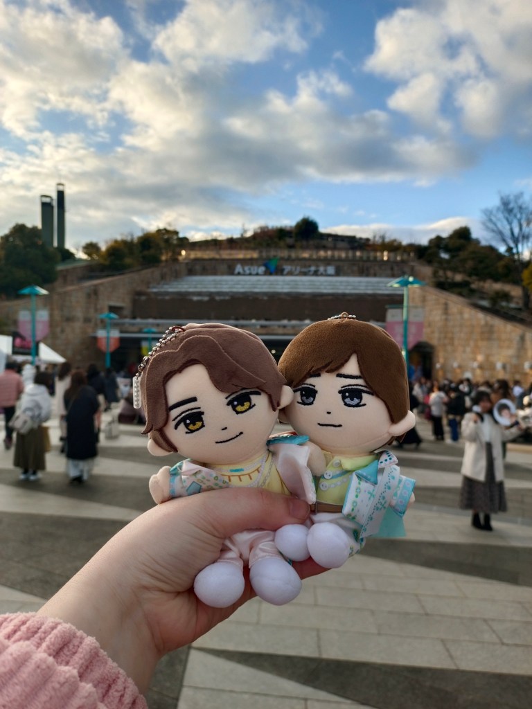 Dos muñecos de peluche que representan a los miembros de King & Prince, sostenidos por una mano, frente al Asue Arena en Osaka con un cielo nublado y un grupo de fans de fondo.