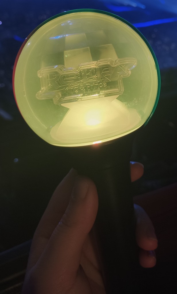 Una mano sostiene un lightstick de color amarillo con el logo de "Re:Era" del grupo musical King & Prince, iluminado en un ambiente de concierto.