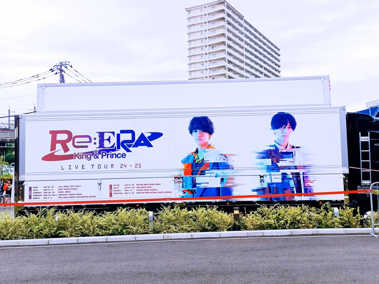 Camión promocional del tour 'Re:ERA' de King & Prince, mostrando a los dos artistas con un diseño gráfico colorido. Se observan detalles de la gira y un fondo urbano.