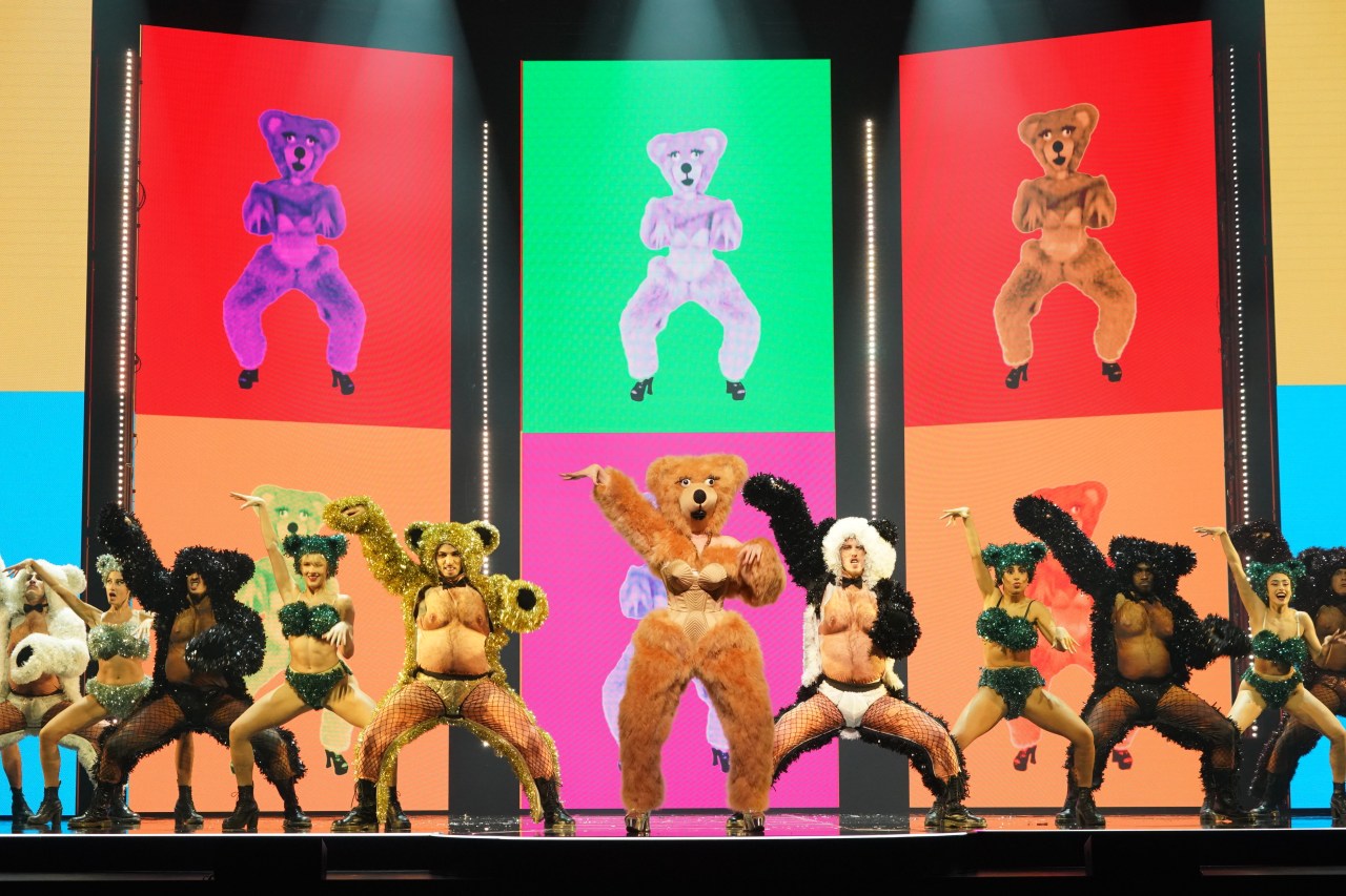 A lively performance featuring dancers in bear-themed costumes, showcasing energetic choreography on stage with colorful, dynamic background visuals.