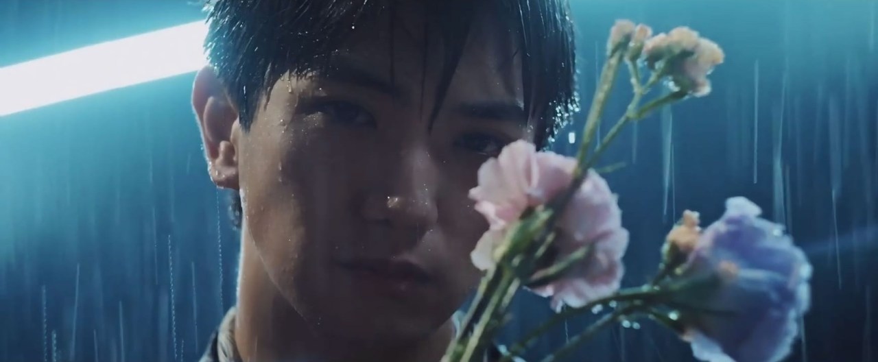 A close-up of a young man holding colorful flowers while standing in the rain, with water droplets glistening on his skin and a soft light illuminating the scene.