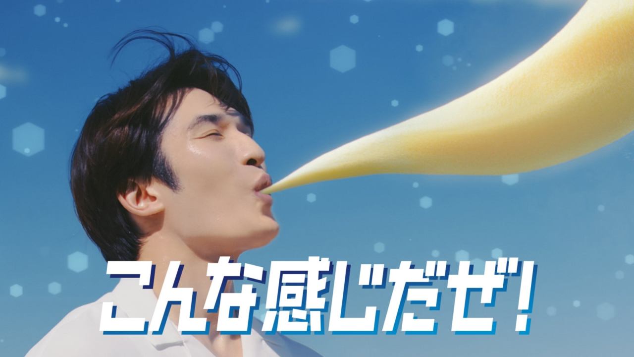 A man with dark hair is smiling while a yellow creamy substance is flowing from his mouth against a clear blue sky, accompanied by the text 'こんな感じだぜ!' in bold white letters.