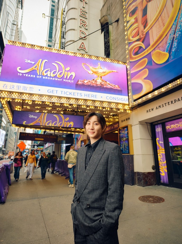A young man in a stylish dark suit stands outside the New Amsterdam Theatre, featuring the 'Aladdin' Broadway display in the background.