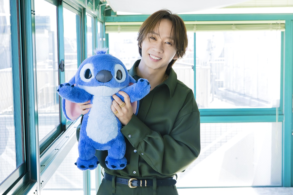 A person with medium-length hair wearing a dark green shirt, smiling while holding a blue Stitch plush toy, standing indoors with large windows in the background.
