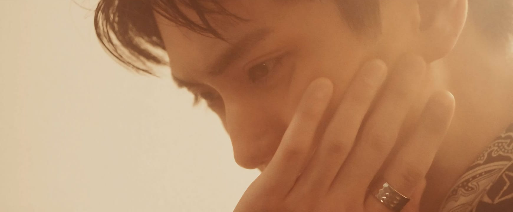 A close-up of a young man's face depicting a moment of contemplation, with his hand gently touching his cheek. The lighting is soft and warm, creating an intimate and reflective atmosphere.