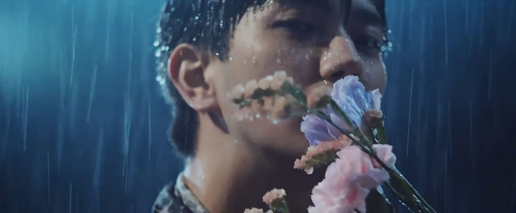 A close-up of a member from Ae! group standing in the rain, holding a bouquet of multicolored flowers close to their face, with water droplets on their skin.