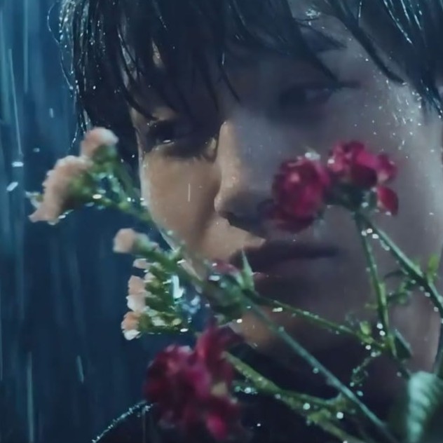 A close-up of a person standing in the rain, holding a bouquet of flowers, with water droplets visible on their face and the flowers.