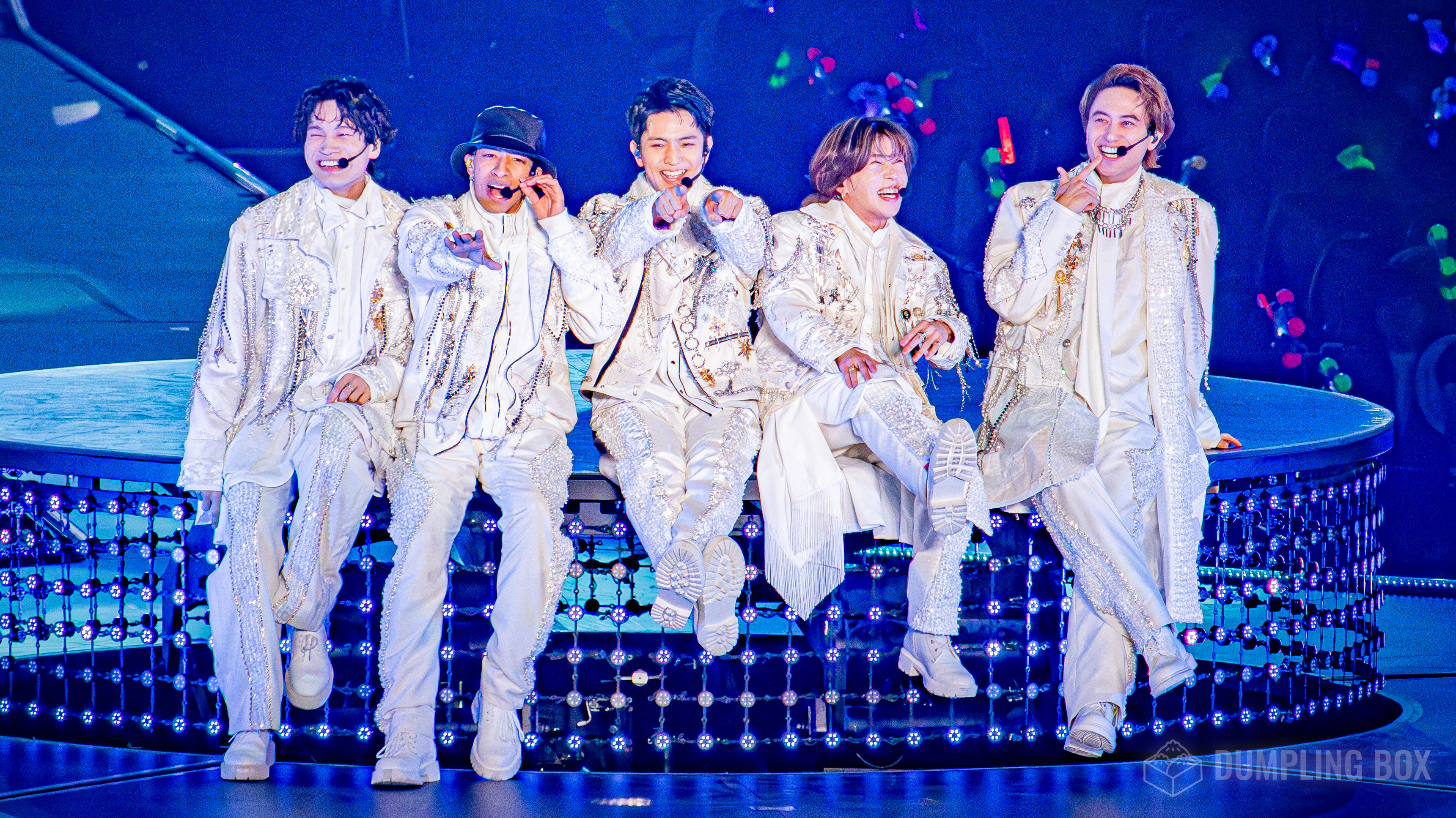 Five members of Ae! group performing together on stage, wearing matching white outfits adorned with sequins, as they share joyful expressions and interact with the audience.