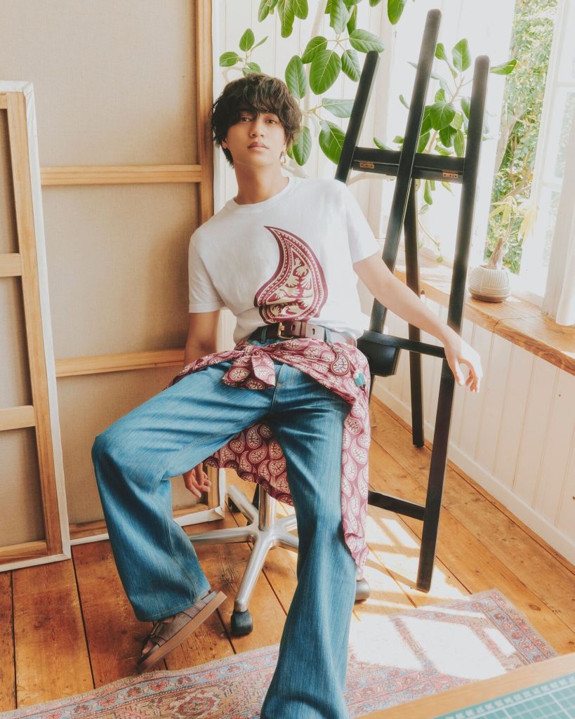 A young man sitting casually in a sunlit room, wearing a white T-shirt with a paisley design and blue jeans. He has a bandana tied around his waist and is posed against a backdrop of wooden furniture and greenery.