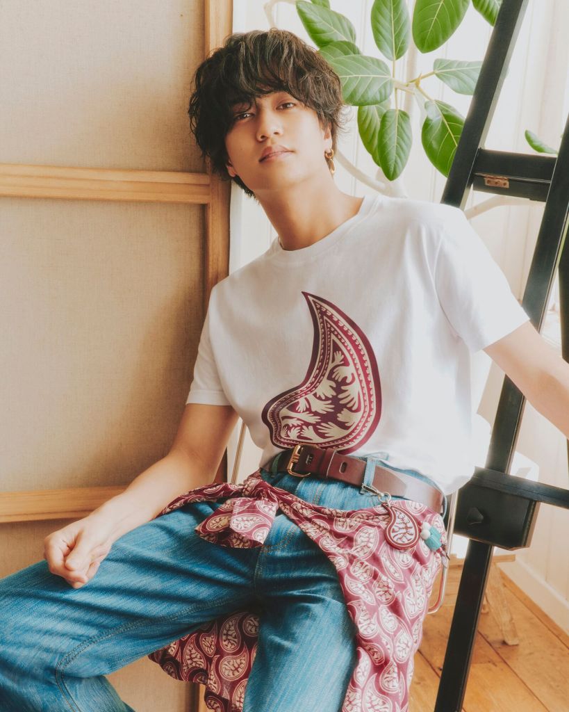 A young man with tousled hair sits casually against a wooden frame, wearing a white t-shirt with a large paisley design. He pairs it with blue jeans and has a patterned cloth draped around his waist. In the background, green plants add a natural touch.