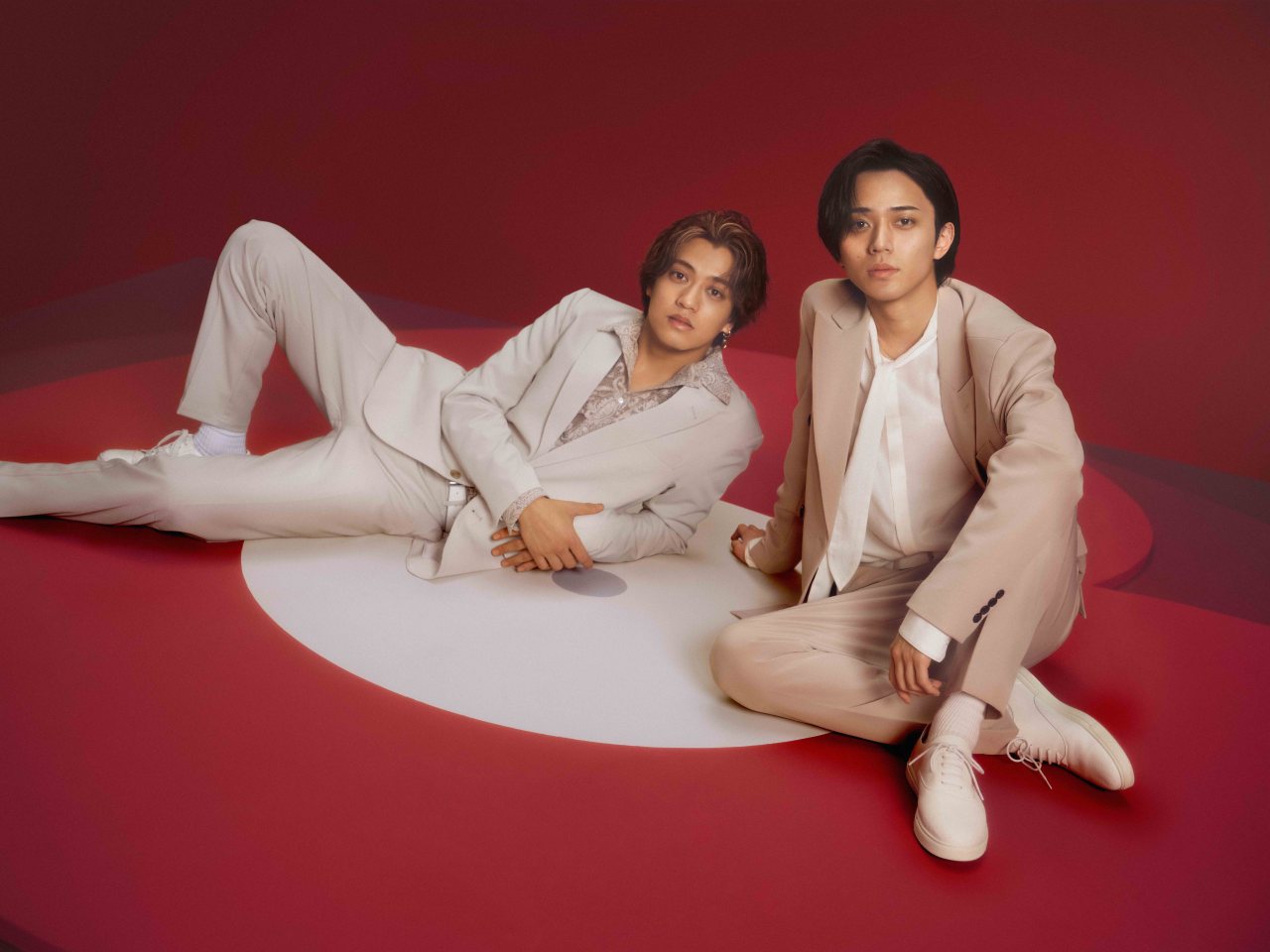 Two young men dressed in light-colored suits are sitting on a colorful circular background, with one leaning back on the floor and the other sitting upright.