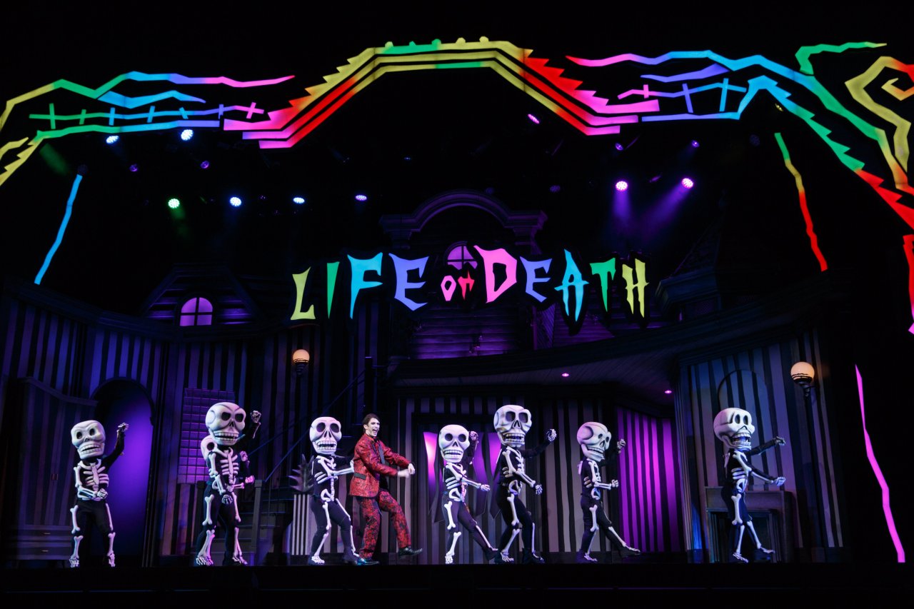 A colorful stage scene from the musical 'Beetlejuice' featuring performers in skeleton costumes dancing, with a vibrant backdrop displaying the words 'LIFE or DEATH'.
