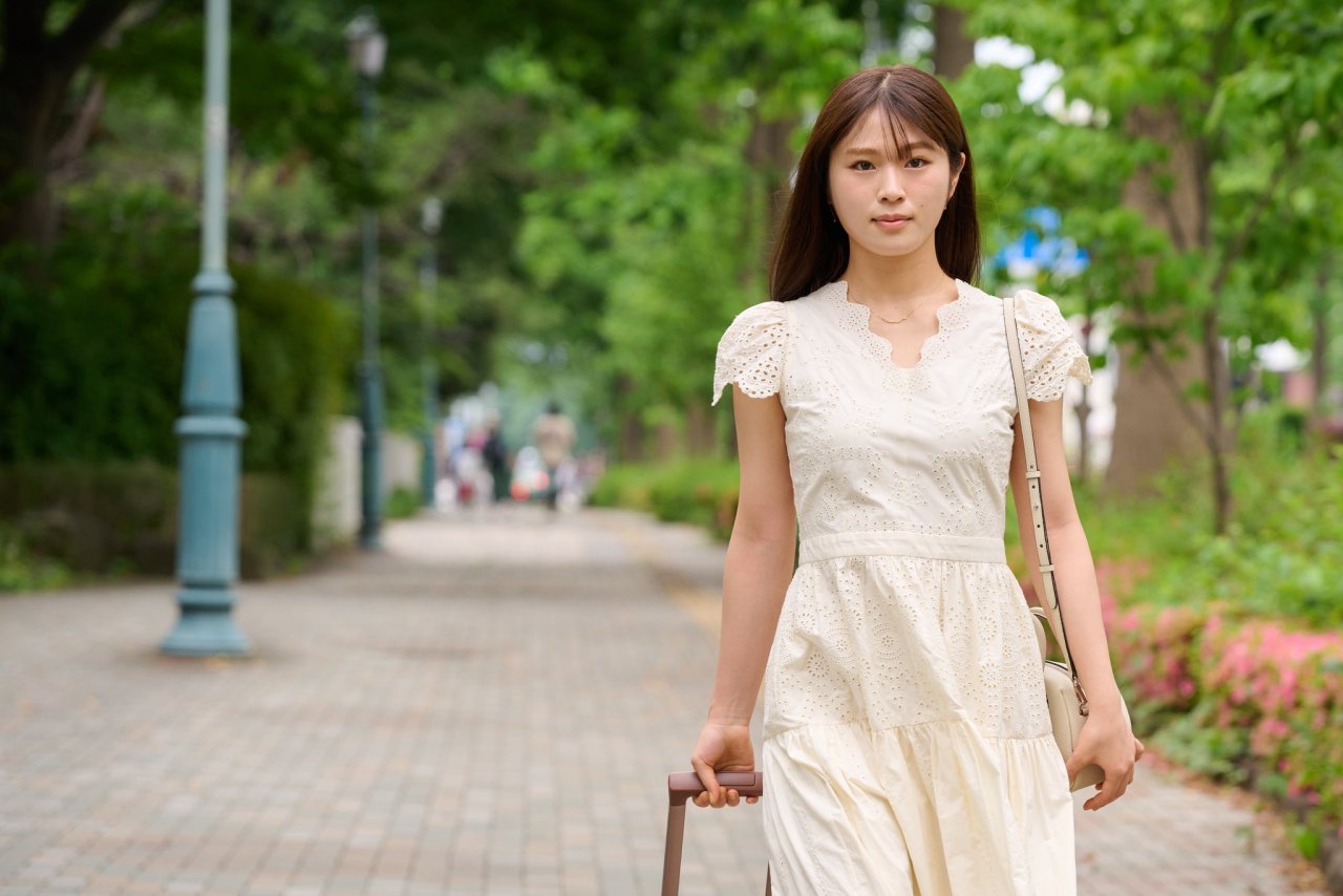 A young woman wearing a white dress walks along a tree-lined pathway, holding a suitcase. She has a serious expression, suggesting a sense of determination.