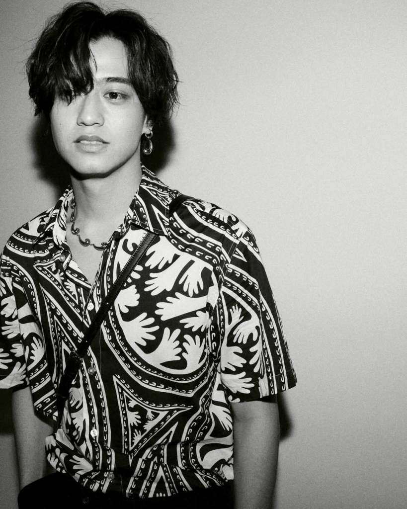 A young man stands against a plain background, wearing a black and white patterned shirt and a necklace, with a stylish haircut and a subtle expression.