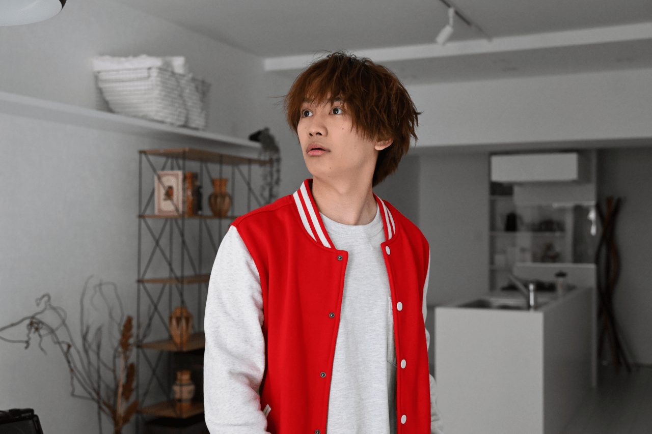 A young man with tousled hair wearing a red varsity jacket and gray shirt, looking surprised in a modern, minimalistic indoor setting.