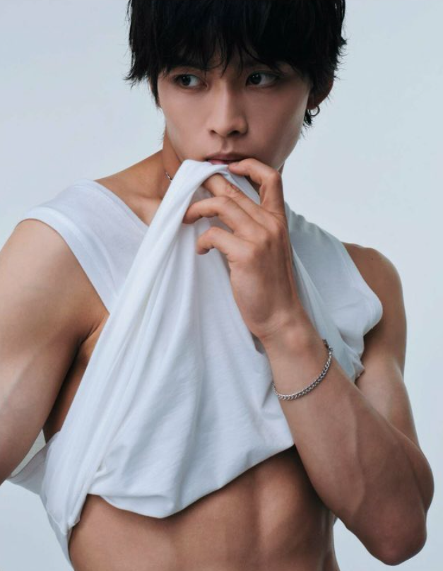 Close-up portrait of a young man with dark hair, wearing a white sleeveless shirt. He holds part of the shirt with his hand near his mouth, showcasing his well-defined abs and a pensive expression.