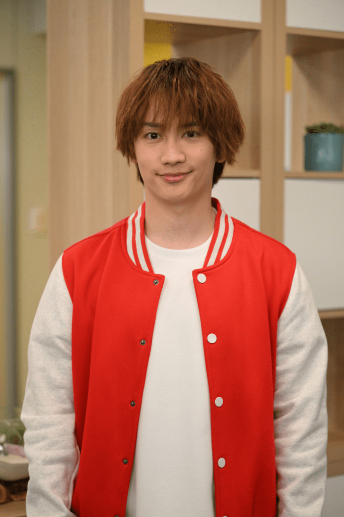 A young male actor smiles while wearing a red bomber jacket over a white shirt, standing in a well-lit room with wooden shelves in the background.