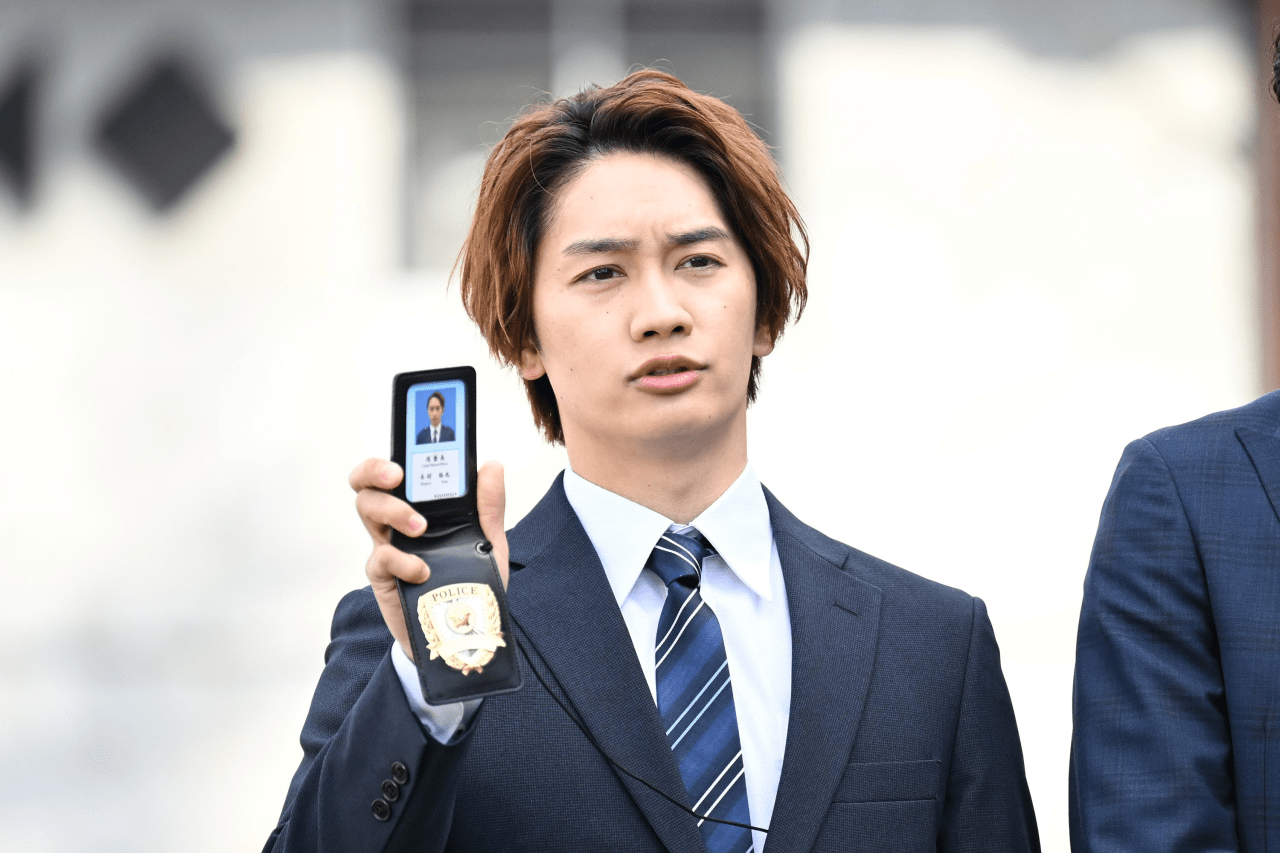 A young man in a suit holds a police badge and looks serious while engaging in a conversation.