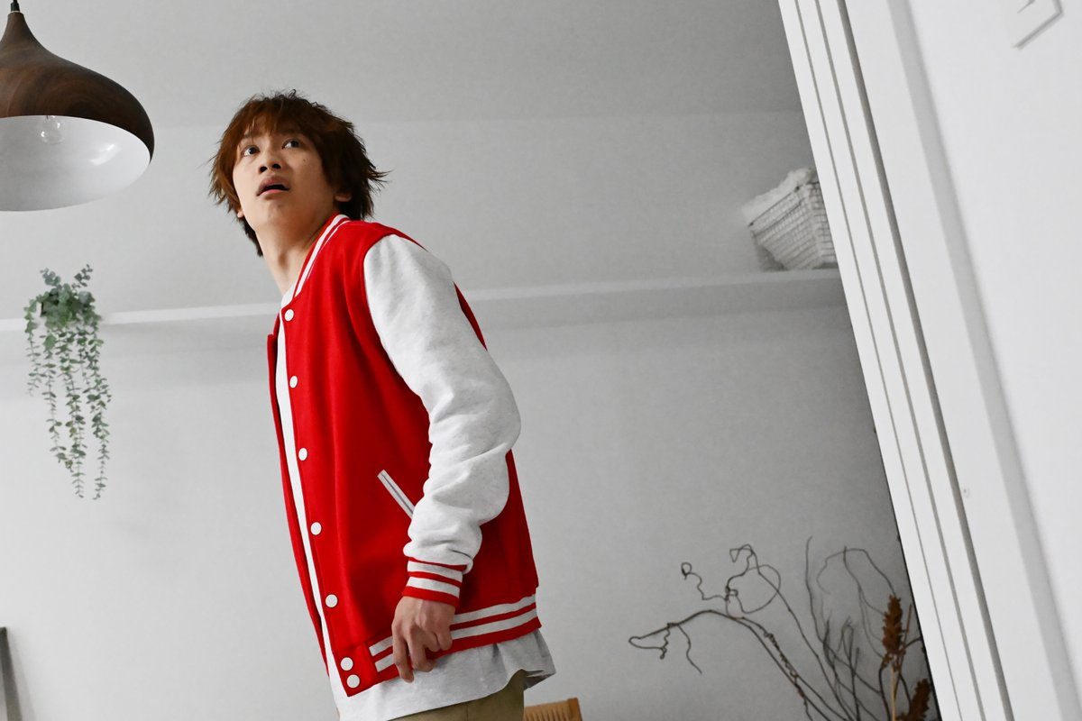 A young man with tousled hair wearing a red varsity jacket looks surprised as he stands in a bright, minimalist room.