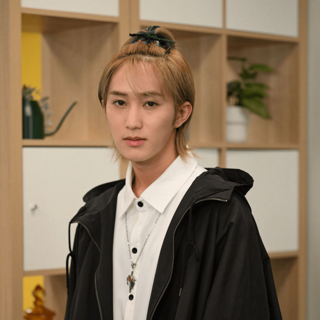 Joichiro Fujiwara posing for a portrait, wearing a black jacket over a white shirt, with a decorative hair clip, set against a backdrop of modern shelving and greenery.