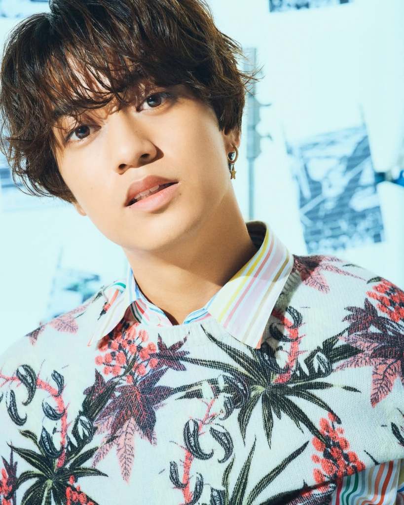 Close-up portrait of a young man with curly hair, wearing a floral patterned sweater and a multi-colored collared shirt against a light background.