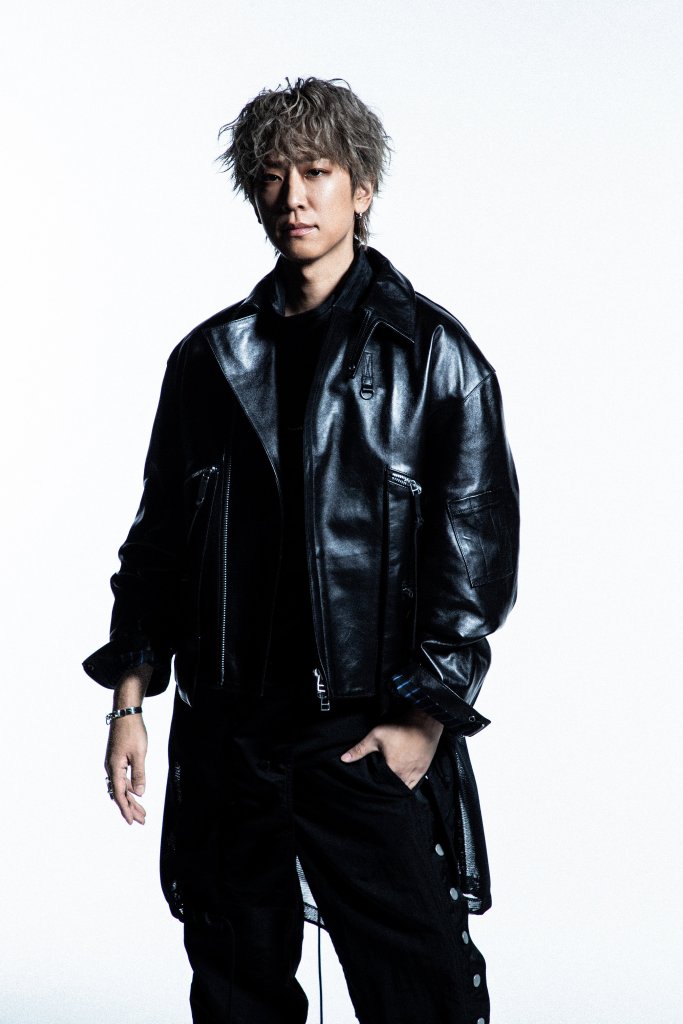 A person with short, spiky hair wearing a black leather jacket over a black shirt, standing confidently against a plain white background.