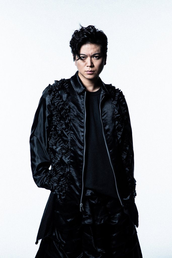 A male artist striking a serious pose, wearing a stylish black jacket with ruffled details over a black top, against a plain white background.