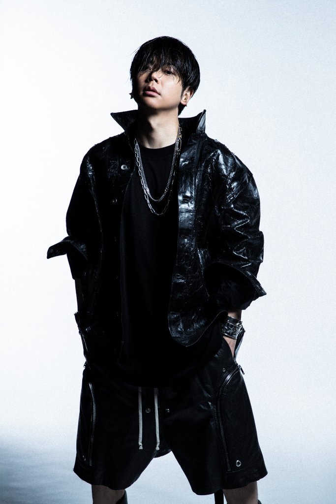 A stylish young man with tousled hair poses confidently in a shiny black oversized jacket and black shorts against a light background.