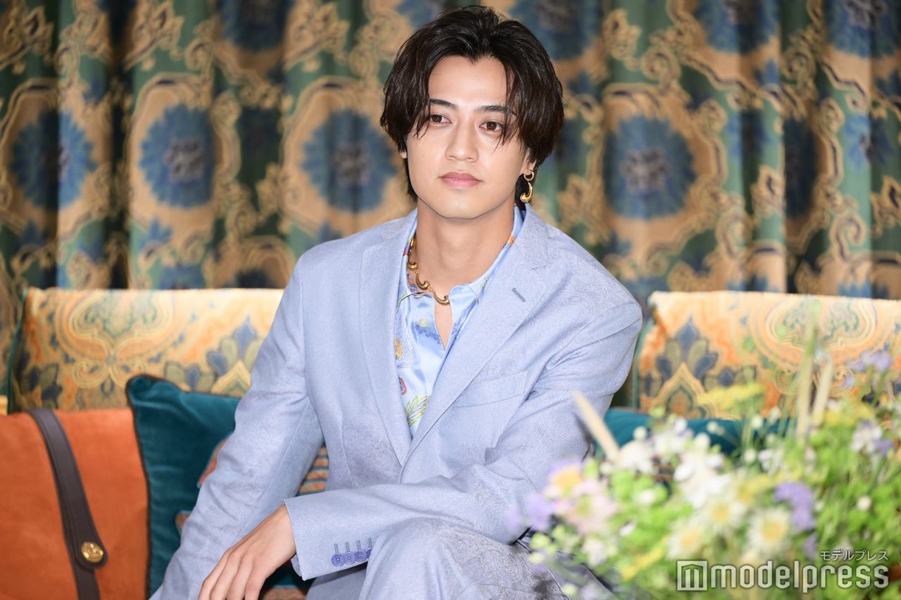 Kaito Takahashi at the Etro Home showroom launch event in Ginza, wearing a light blue suit and a colorful shirt, seated with decorative fabric and flowers in the background.