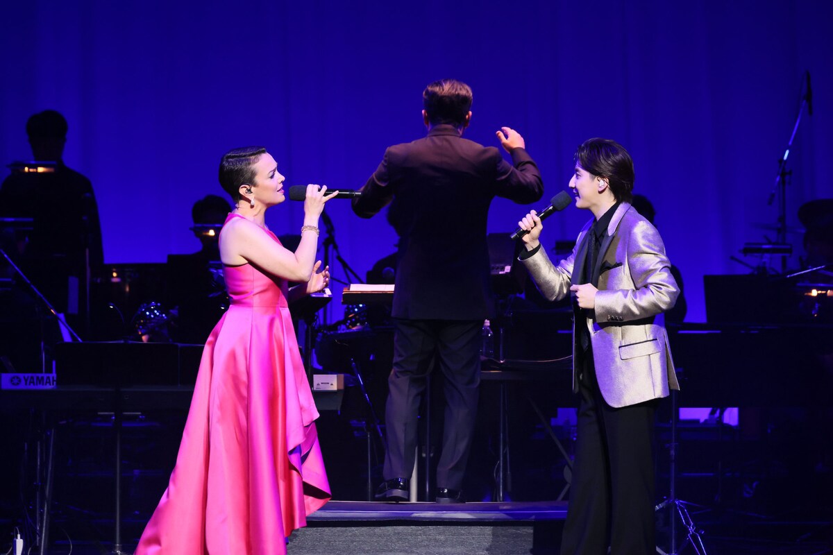 A musical performance featuring a female singer in a pink dress and a male singer in a silver jacket, both holding microphones, while a conductor is in the background, surrounded by a band in a dimly lit venue.