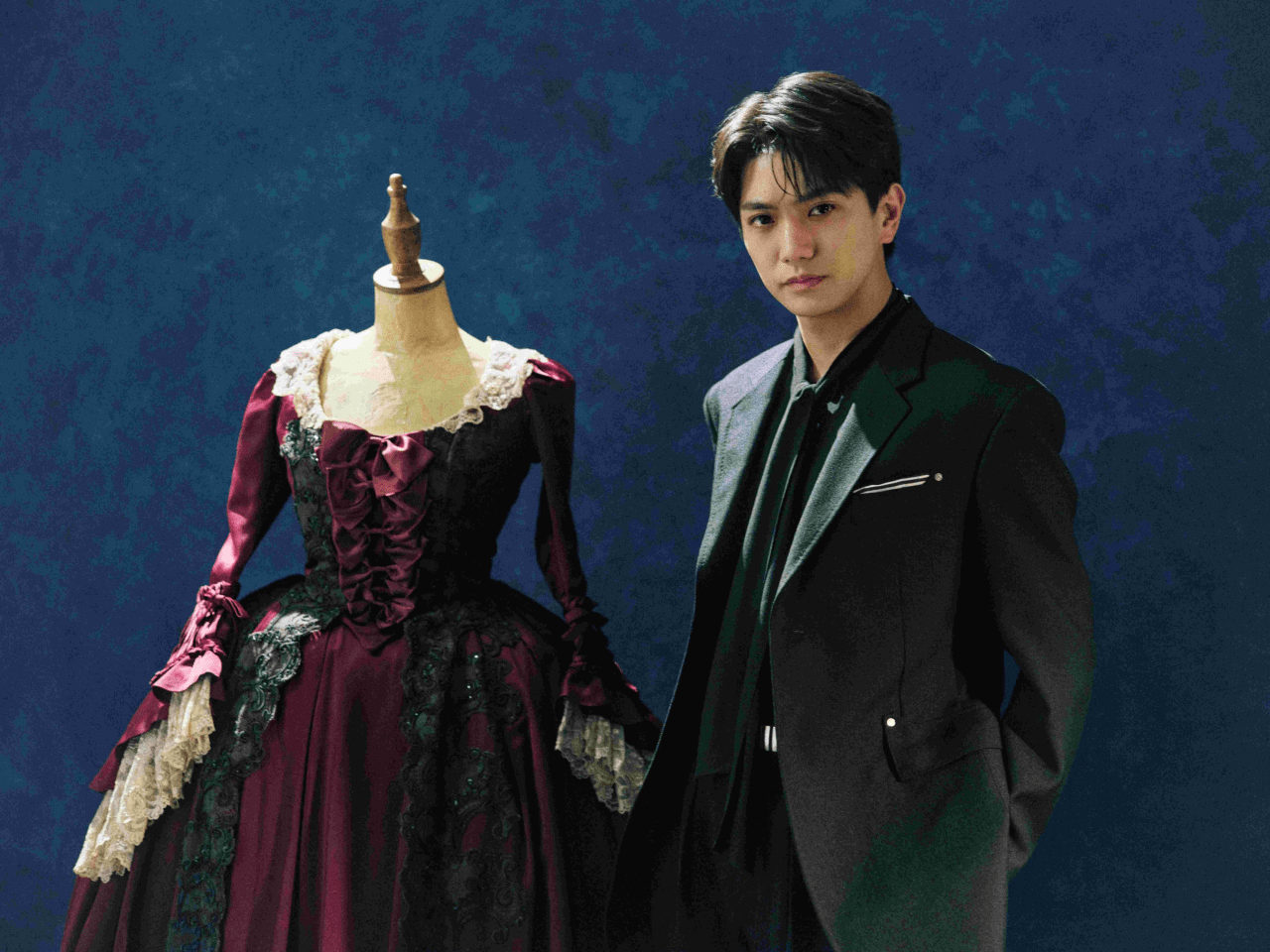 Yoshinori Masakado poses alongside a vintage dress on a mannequin, wearing a black suit against a blue background.