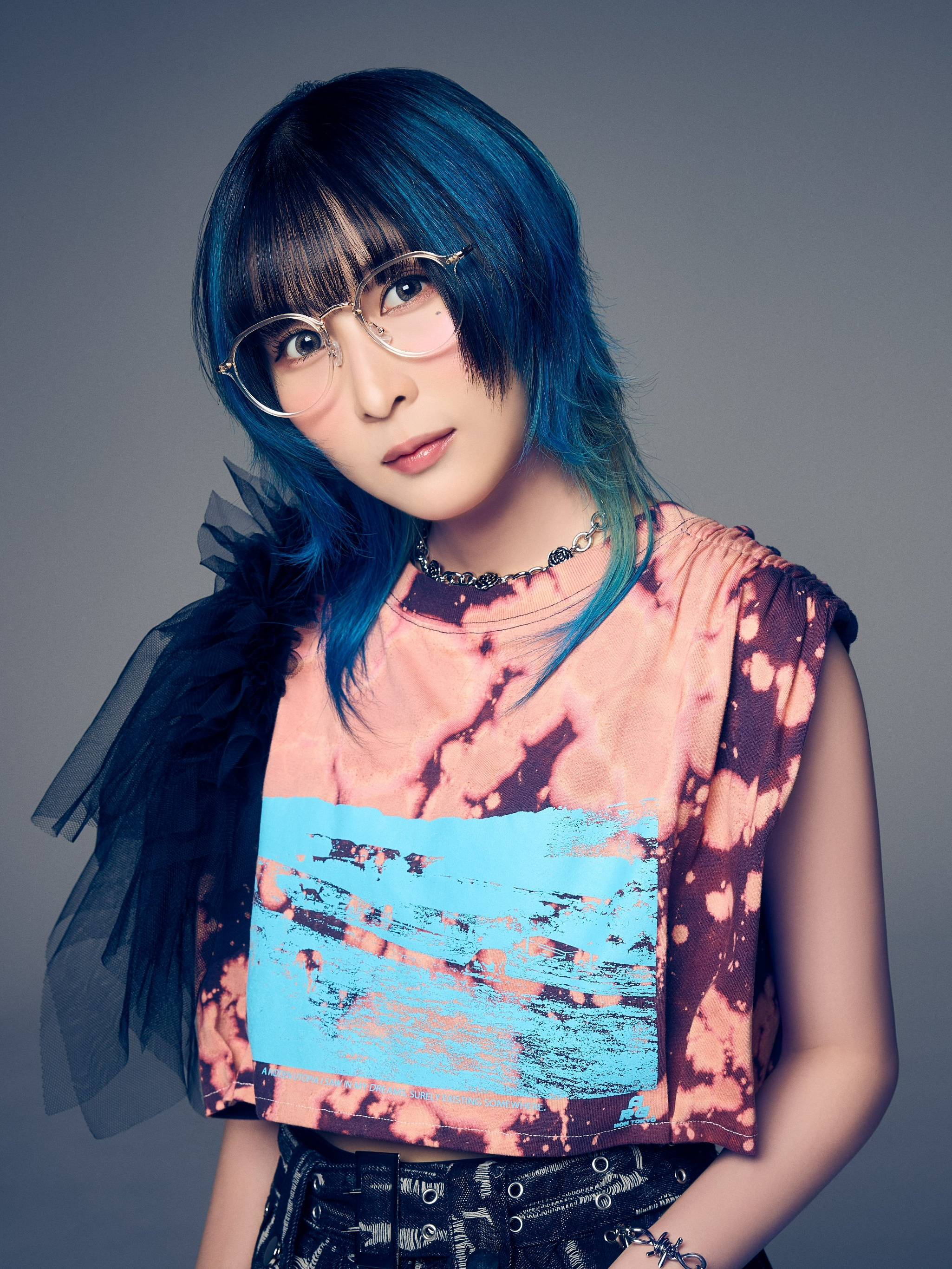 A close-up portrait of MindaRyn, a Thai anisong singer, featuring her with blue-tinted hair and stylish glasses, wearing a vibrant tie-dye shirt and accessories.