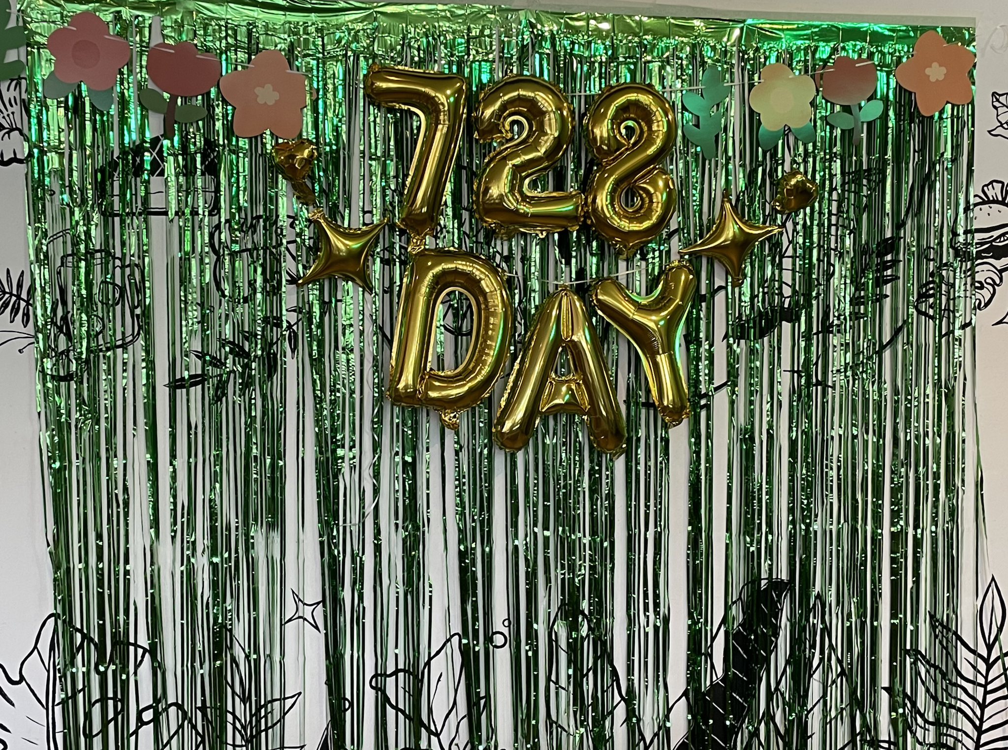 A decorative display featuring golden balloons spelling '728 DAY' against a backdrop of shiny green streamers and floral cutouts.