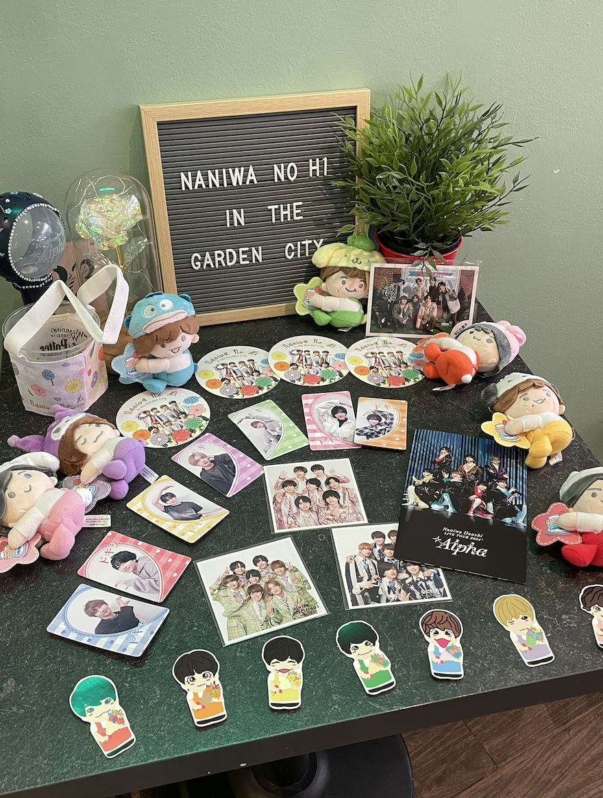A table display filled with various Naniwa Danshi themed merchandise, including plush toys, stickers, and photo prints, alongside a decorative sign reading 'NANIWA NO HI IN THE GARDEN CITY'.