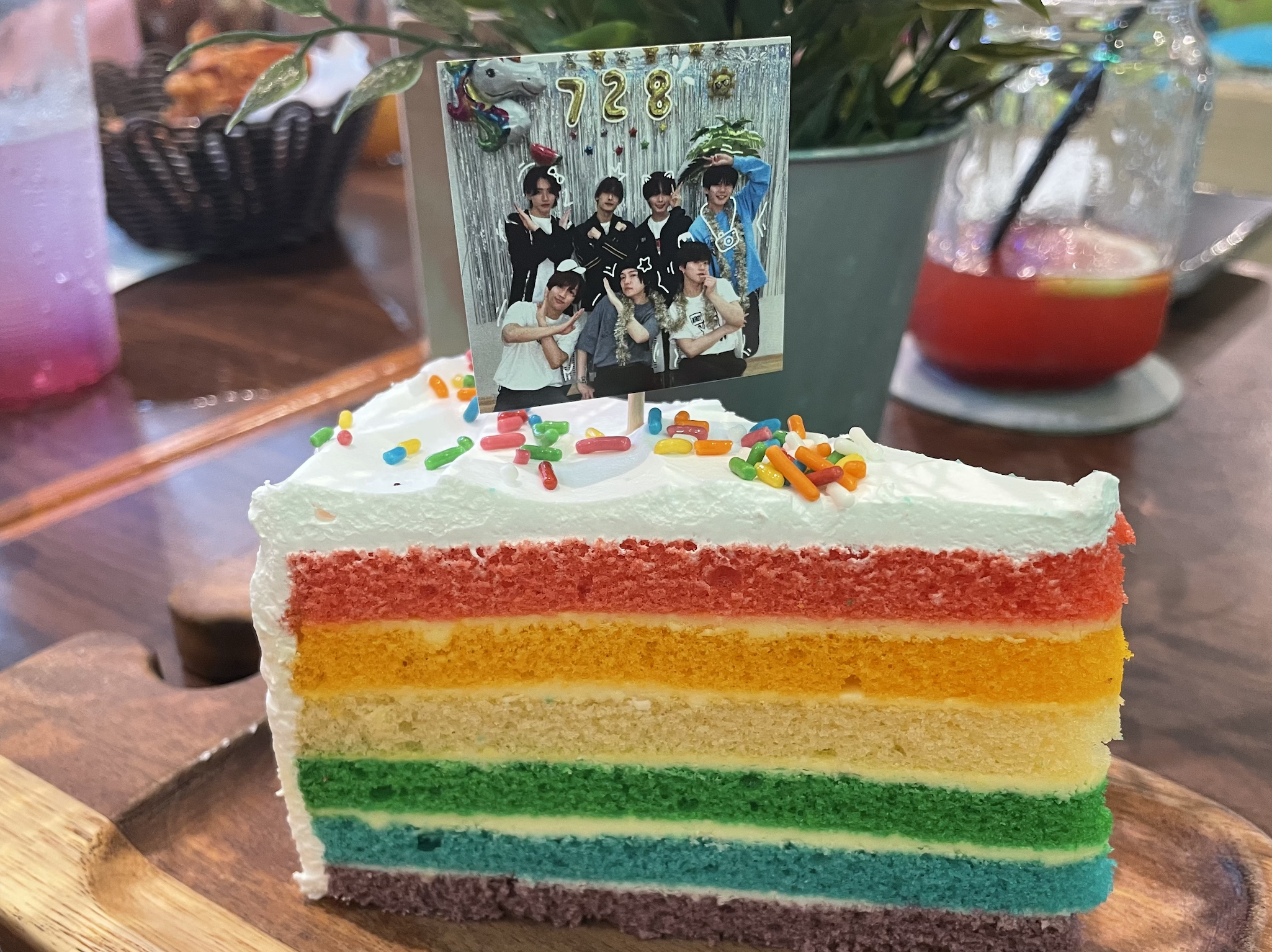 A colorful rainbow cake slice topped with whipped cream, rainbow sprinkles, and a photo of seven individuals, displayed on a wooden plate in a cafe setting.