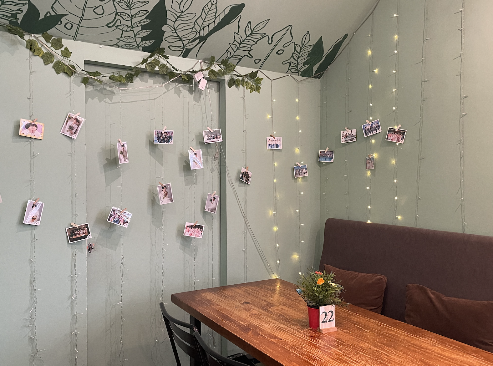 A cozy cafe interior decorated with fairy lights and photos hanging on strings, featuring a wooden table with a small potted plant and a table number.