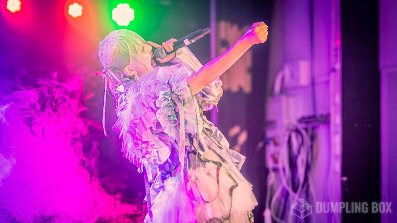A performer with light green hair sings passionately into a microphone, wearing a unique, ruffled outfit. Colorful stage lights in the background create an energetic atmosphere, with pink and green hues visible.