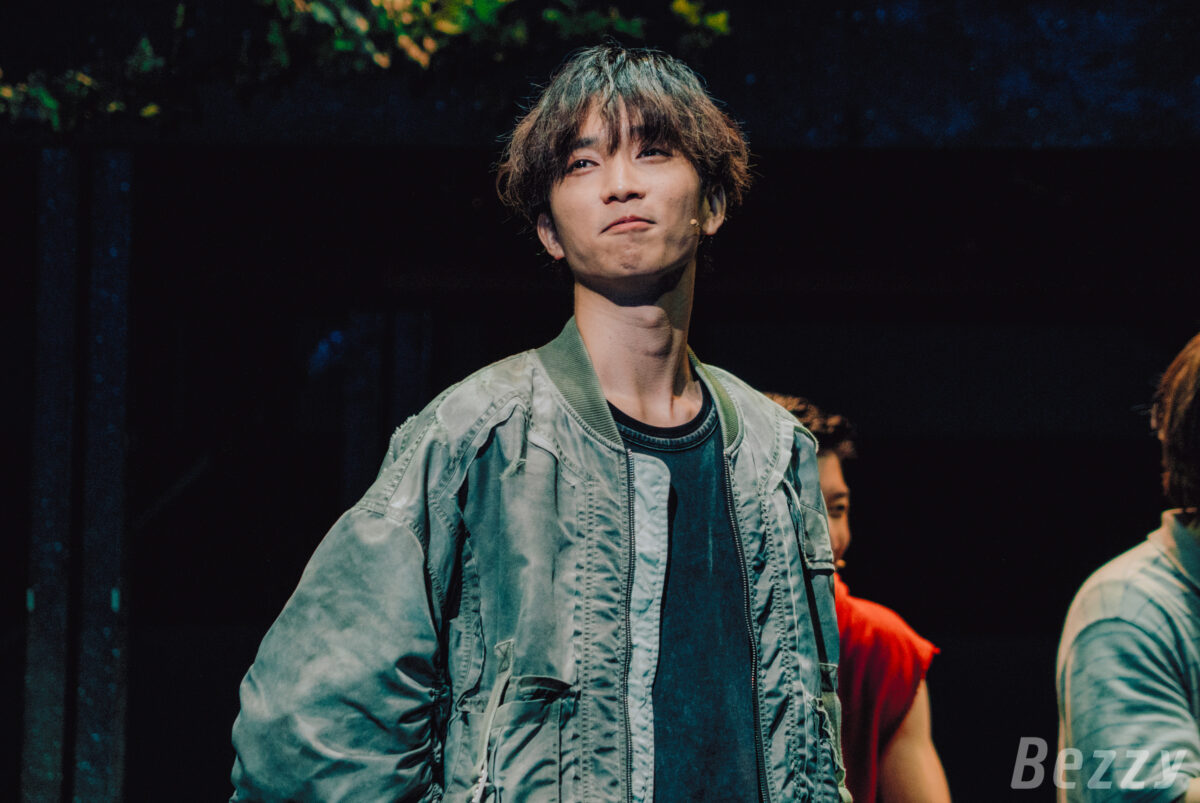 Juri Tanaka performing on stage in the play 'Bokura no nanoka kan Sensou 2025,' wearing a green bomber jacket and a serious expression.