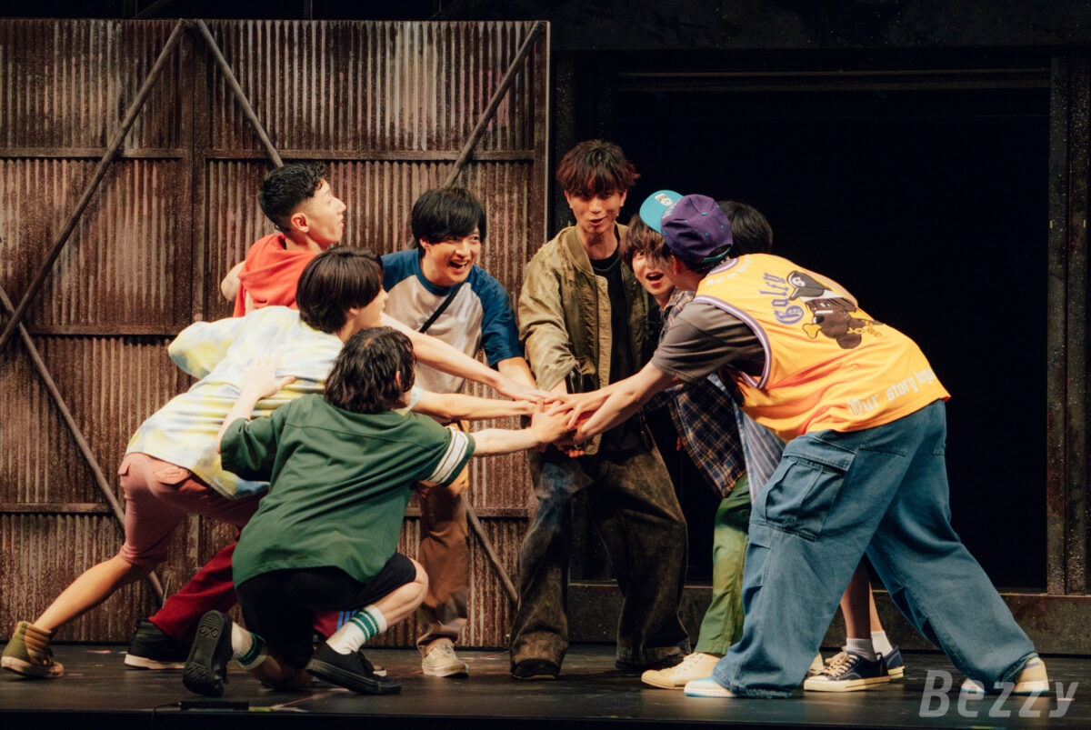 A group of young male actors wearing casual clothing huddle together on stage, playing a game or engaging in a moment of camaraderie, with a metal backdrop creating an industrial atmosphere.