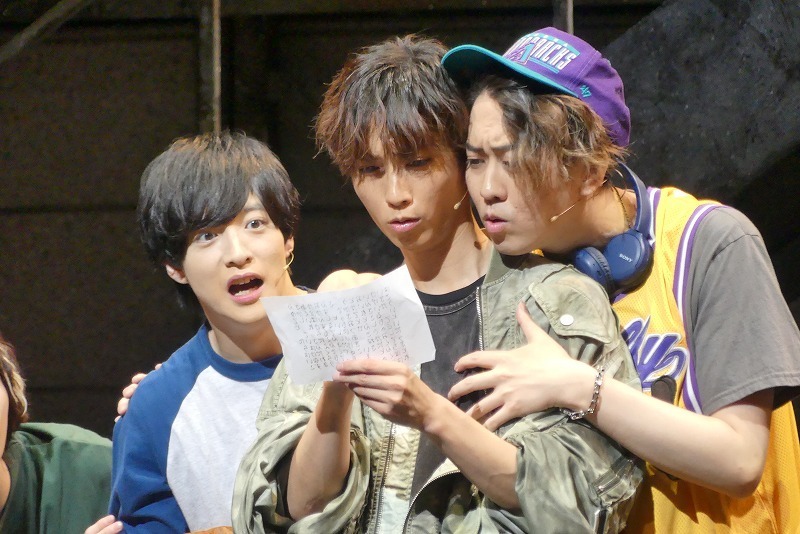 Three young male actors on stage are intently reading a piece of paper, showing expressions of surprise and concern.