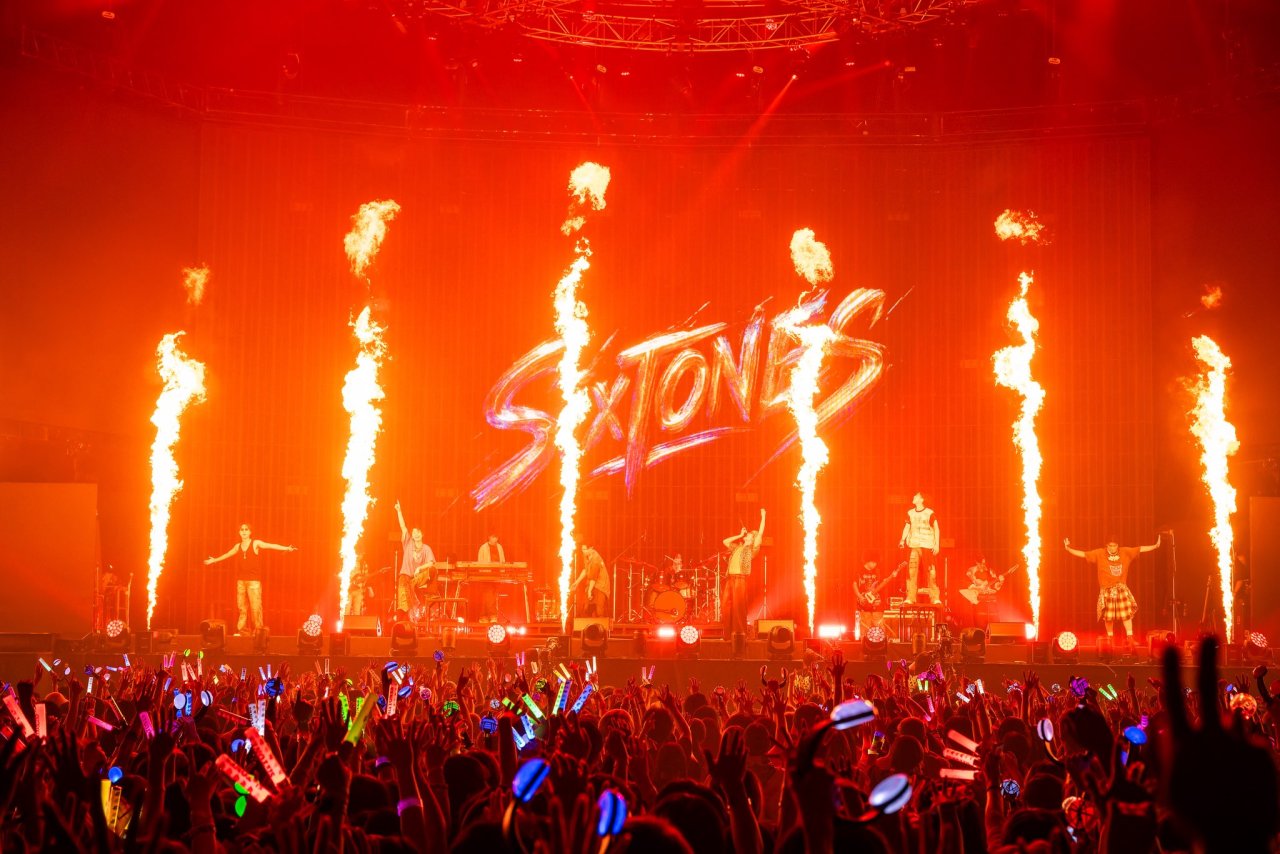A vibrant live performance by the group SixTONES, with colorful lights and flames on stage, engaging a cheering crowd holding glow sticks.