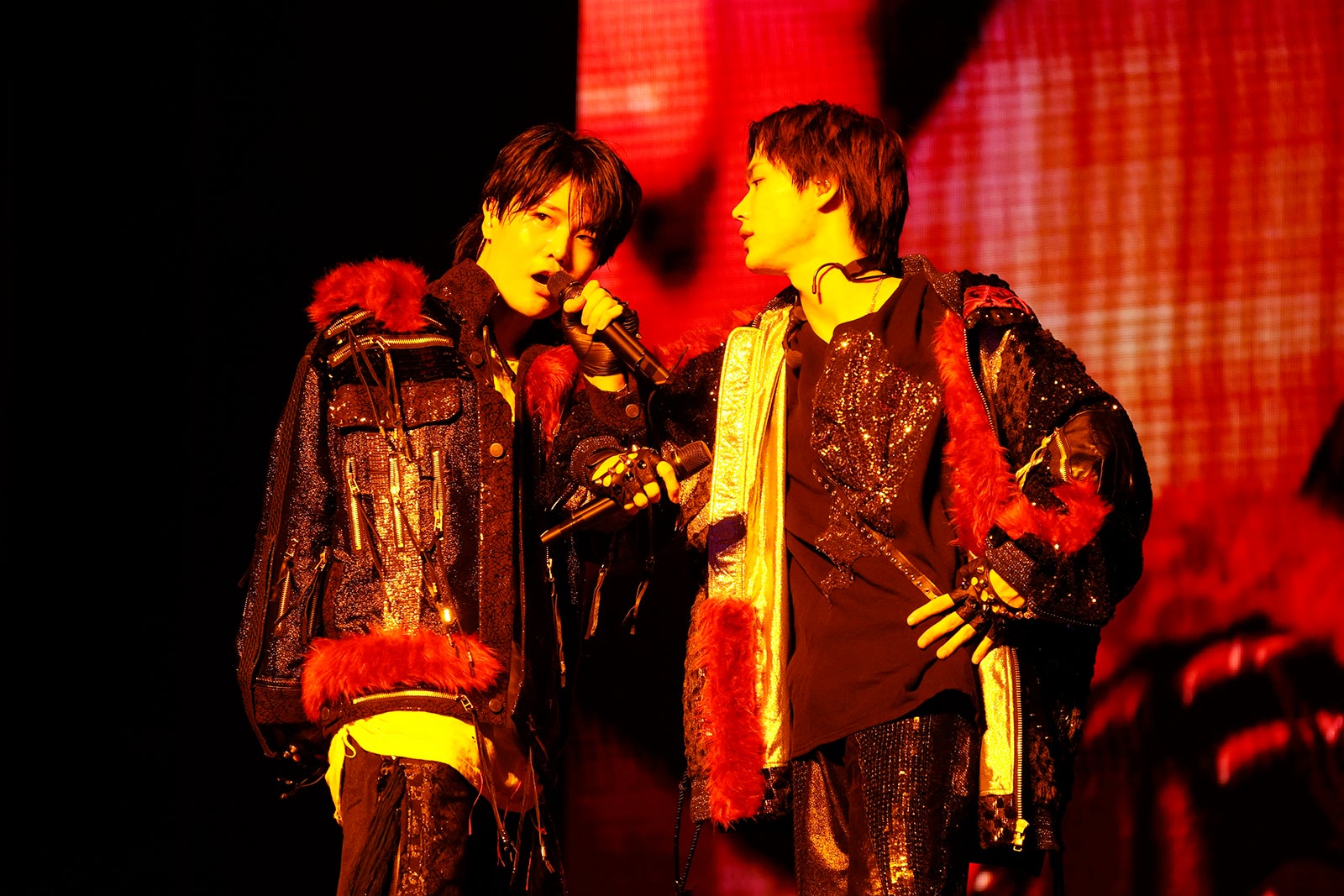 Two performers in extravagant stage costumes engage in a dynamic duet on stage, surrounded by vibrant lighting and visual effects.