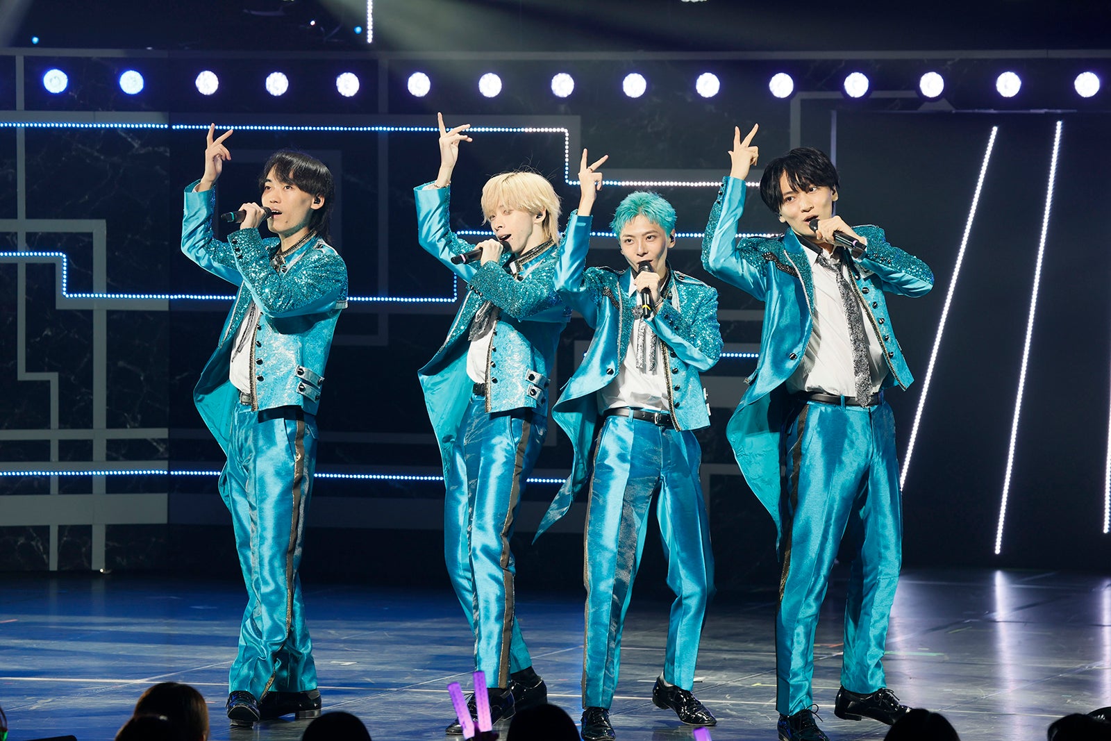 Four performers dressed in sparkling turquoise suits are singing on stage, each holding a microphone and gesturing energetically during a concert.
