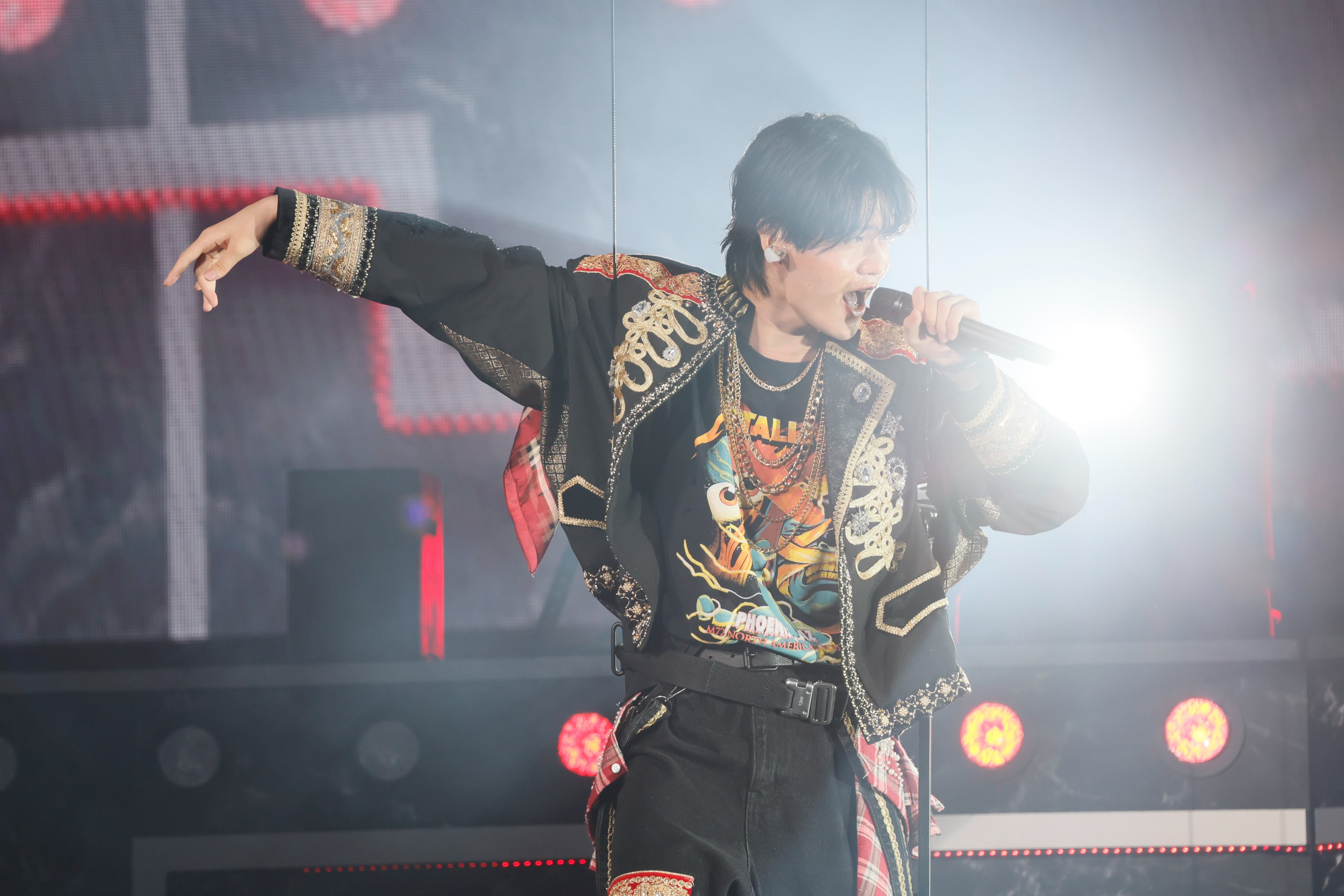 A performer dressed in a stylish black and gold outfit sings passionately into a microphone on stage, surrounded by bright stage lights and a vibrant atmosphere.