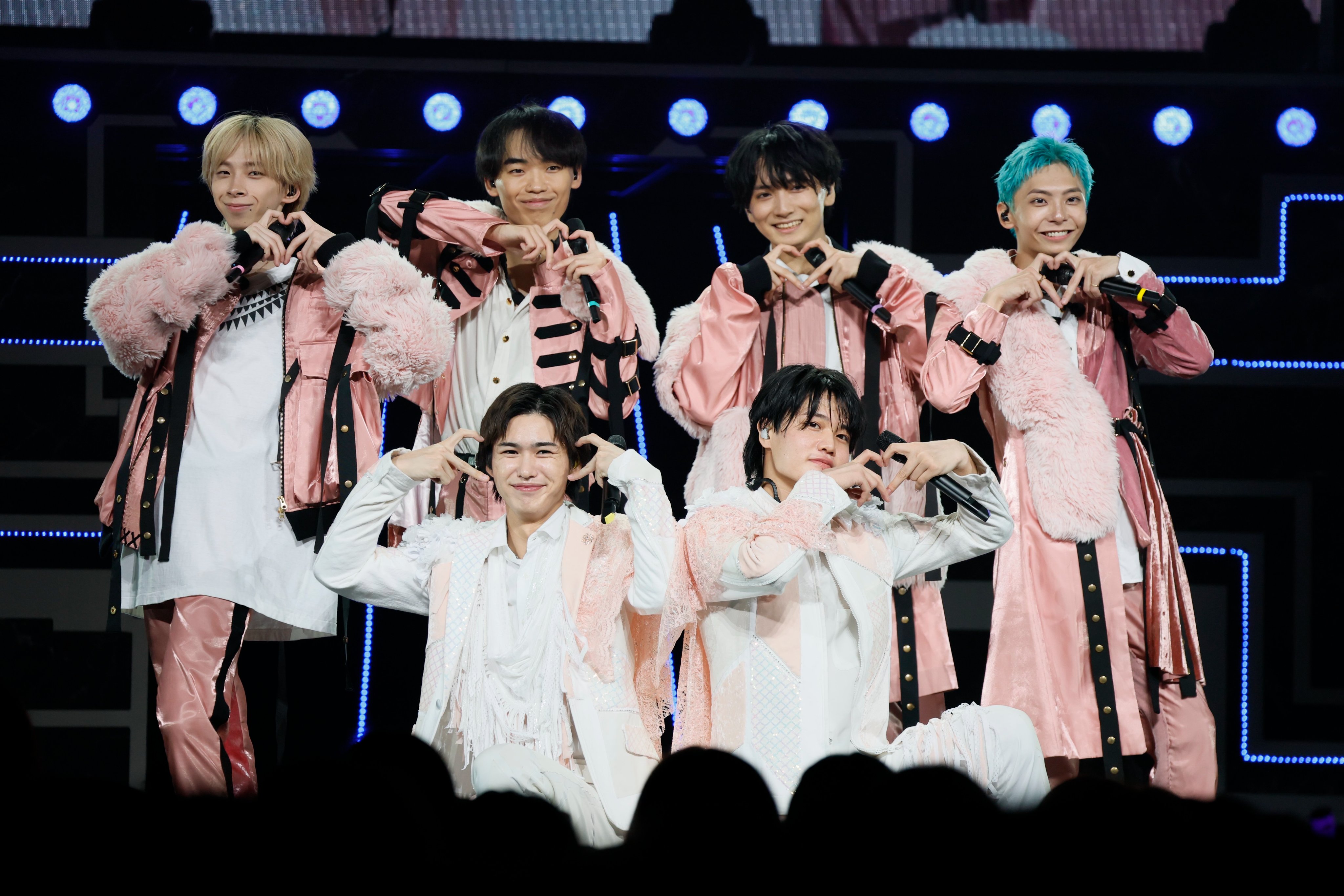 A group of six performers on stage, dressed in stylish pink outfits, posing with heart shapes made with their hands.