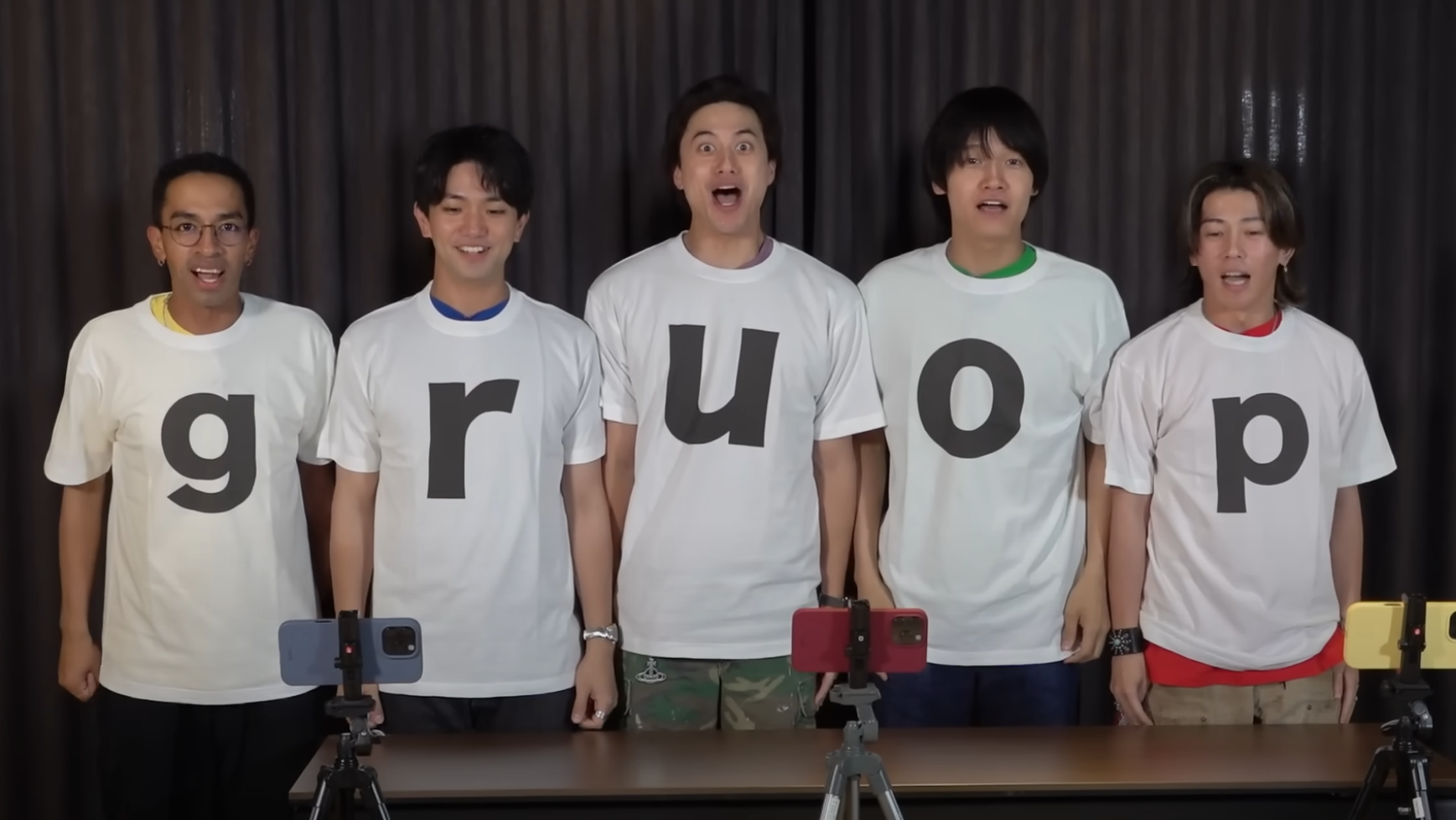 Five members of Ae! group smiling and wearing white t-shirts with large black letters spelling 'group,' standing in front of cameras on a table.