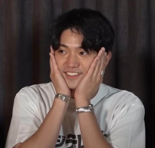 A member of Ae! group smiling with hands on their cheeks in a playful expression, wearing a light-colored t-shirt and sitting against a dark backdrop.