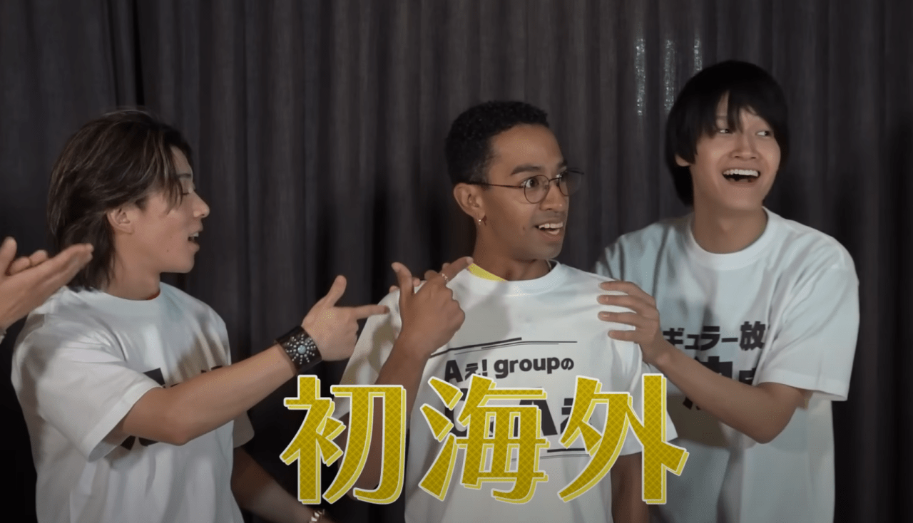 Members of Ae! group expressing excitement and surprise, dressed in matching T-shirts, with a decorative backdrop highlighting the word 'ๆตทๅค' (overseas).